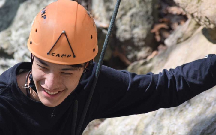 A person wearing a helmet and secured by a rope smiles as they appear to rock climb. 